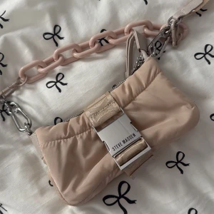 Beige axelväska från Steve Madden - Trendig beige axelväska från Steve Madden med bred axelrem, chunky plastkedja i ljusrosa och coolt spänne framtill. Väskan är gjord i mjukt syntetmaterial och har dragkedja upptill samt innerfack. 