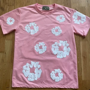 Rosa DenimTears tshirt  - Säljer en rosa t-shirt från Cav Empt med unika vita grafiska tryck i cirkelform över hela tröjan. Klassisk passform med rund hals och korta ärmar. Tillverkad i mjuk bomull som känns skön mot huden. Perfekt för dig som gillar streetwear med en twist.