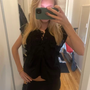 Svart blus med snörning från Neo Noir - Supersnygg svart blus från Neo Noir med volangdetaljer och snörning framtill. Blusen har korta ärmar och en luftig passform, perfekt för att lyfta vilken outfit som helst. Tillverkad i mjuk bomull som känns skön mot huden.