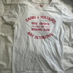 Vit Zadig & Voltaire t-shirt med tryck - Vit t-shirt från Zadig & Voltaire med rött tryck på bröstet där det står 'ART IS TRUTH' och 'RIVE DROITE'. Klassisk rund hals och korta ärmar. Skön och lätt i bomull, perfekt till jeans eller shorts.