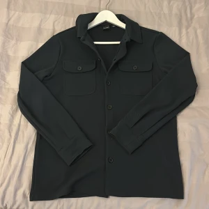 Svart overshirt från Only & Sons - Stilren svart overshirt från Only & Sons med klassisk krage, två bröstfickor med knappar och långa ärmar. Skjortan har en clean look och är tillverkad i ett mjukt, lite stretchigt material som känns bekvämt. Perfekt att bära öppen eller stängd. Använd enstaka gånger! Passar även S!