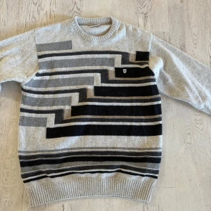 Grå stickad tröja med grafiskt mönster - Snygg grå stickad tröja med svart och mörkgrått grafiskt mönster framtill. Tröjan har rund halsringning, ribbade muddar och långa ärmar. Perfekt för dig som gillar retrovibbar och vill ha något unikt i garderoben.