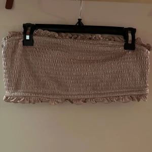 Beige bandeau tubtopp från H&M strl: 46 - Supersöt beige bandeau tubtopp från H&M med smockad struktur och volangdetaljer upptill och nedtill. Toppen är ärmlös och har en mjuk, stretchig känsla tack vare materialmixen av polyamid, polyester och elastan. Perfekt för en trendig och avslappnad look.