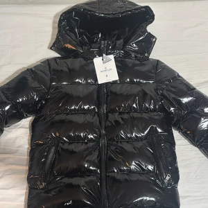 Svart glansig dunjacka från Moncler - Säljer en svart, glansig dunjacka från Moncler med huva och dragkedja framtill. Jackan har två fickor med dragkedja, quiltad design och klassisk Moncler-logga på ärmen. Perfekt för kalla dagar och riktigt snygg streetstil. Helt ny aldrig använd 