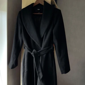 Svart lång kappa - Supersnygg och stilren svart kappa från Bik Bok med knappar och knytskärp i midjan!!🤎🤎 Använd mycket sparsamt, inga defekter!☺️Perfekt till hösten! Nypris 900 kr