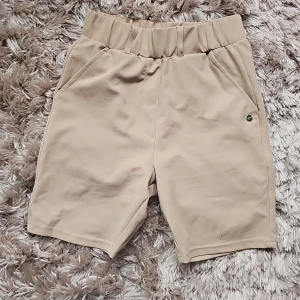 Beiga shorts från Zink i S - Stilrena beiga shorts/cykelbyxor från Zink med elastisk midja och bälteshällor. Modellen har fickor fram och dekorativa fickor bak. Mjukt tyg med lätt struktur, perfekt för varma dagar. Klassisk och enkel design som är lätt att matcha.
