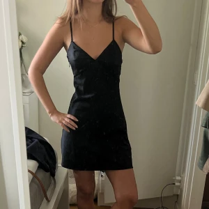 Svart satin klänning med smala axelband - Säljer en svart kort satin klänning med smala axelband och v-ringning. Klänningen har en figurnära passform och är perfekt för dig som gillar en enkel men stilren look. Materialet är slätt och ger en snygg siluett. Passar dig som vill ha en klassisk och tidlös stil.