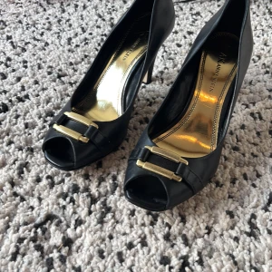 Svarta pumps från Anne Klein med gulddetalj - Stilrena svarta pumps från Anne Klein i skinn med öppen tå och snygg guldfärgad detalj framtill. Skorna har klack och en elegant form som ger en klassisk look. Perfekta för dig som vill ha något extra till outfiten. Bra för både fest men oxå en stilren look. Kontakta vid frågor, pris kan diskuteras då jag vill få dem sålda! Storleken motsvarar ca 38