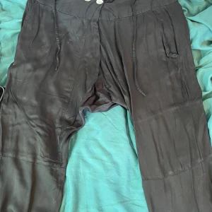 Svarta loose byxor med snörning - Svarta byxor med avslappnad passform och snörning i midjan. De har fickor på sidorna och är tillverkade i ett mjukt, tunt material som känns svalt mot huden. Perfekta för en chill och bekväm stil.