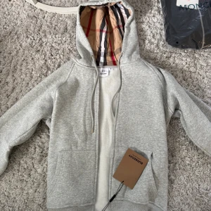 Grå hoodie från Burberry med dragkedja - Snygg grå hoodie från Burberry med klassiskt rutigt foder i huvan. Tröjan har dragkedja framtill, snörning vid huvan och två fickor. Perfekt för dig som gillar stilrena och exklusiva plagg med en twist.