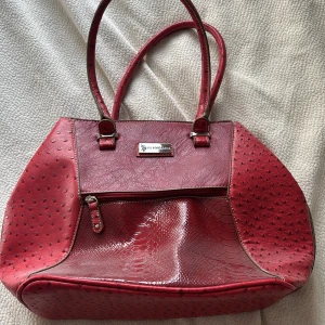 Röd handväska från U.S. Polo Assn. - Snygg röd handväska från U.S. Polo Assn. med två långa handtag och coola detaljer i olika strukturer, som ormskinnsmönster och prickig textur. Väskan har dragkedja framtill och silverfärgade metalldetaljer. Perfekt för dig som vill sticka ut. Köpt för 1200