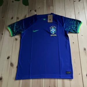 Blå Brasilien fotbollströja Nike M - Snygg blå Brasilien fotbollströja från Nike med grönt Swoosh och CBF-emblem på bröstet. Ärmarna har leopardmönster i blått och grönt med gula muddar. Tillverkad i lätt Dri-FIT material som andas. Perfekt för dig som älskar landslagsfotboll.
