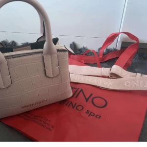 ROSA  handväska från Valentino - Snygg babypink  handväska från Valentino med croco-mönstrad yta och guldfärgad logga. Väskan har både handtag och en bred axelrem med Valentino-tryck, samt dragkedja upptill. Perfekt storlek för det viktigaste och riktigt trendig design.