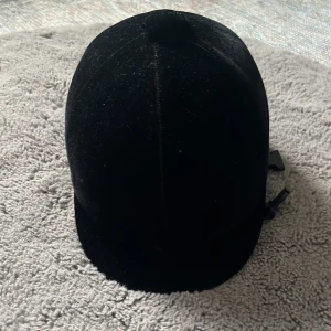 Svart sammet rid hjälm stl 57 - Säljer en klassisk svart ridhjälm i sammet med rundad form och liten skärm framtill. Hjälmen har en dekorativ knapp på toppen och blått innerfoder. Perfekt för dig som vill ha en stilren look i stallet. Aldrig använt för stor för mig 