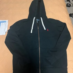 Svart hoodie från Polo Ralph Lauren - Svart hoodie från Polo Ralph Lauren med dragkedja och vit snörning i huvan. Klassisk röd logga broderad på bröstet. Tillverkad i mjuk bomullsblandning med polyester. Perfekt för chill dagar och snygg streetstil. Storlek L men passar som M!