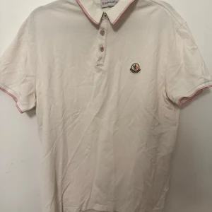 Vit Moncler pikétröja med rosa detaljer - Vit pikétröja från Moncler med klassisk krage, tre knappar och rosa detaljer på krage och ärmslut. Broderad Moncler-logga på bröstet. Tillverkad i bomull för en skön och luftig känsla. Perfekt för dig som gillar stilrena och exklusiva plagg. Står xl men sitter som s