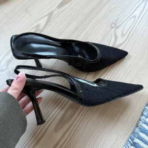 Svarta spetsiga pumps med klack - Säljer ett par svarta pumps med spetsig tå och öppen häl. Skorna har hög klack och är täckta med ett snyggt spetsliknande tyg och blanka detaljer runt kanterna. Perfekta för dig som vill ha en elegant och trendig look. Storlek 38