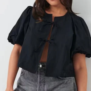 Svart blus med puffärm och knyt - Svart blus med korta puffärmar och trendiga knytband framtill. Modellen är croppad och har rund halsringning. Perfekt för dig som gillar statement-ärmar och en cool, avslappnad look. Tillverkad i bomull för en skön känsla. 
