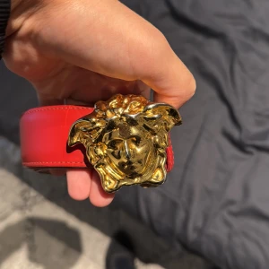 Rött Versace bälte med Medusa-spänne - Säljer ett snyggt rött Versace bälte i äkta skinn med ikoniskt Medusa-spänne i guld. Bältet är brett och har exklusiv känsla, perfekt för att lyfta din outfit. Kommer med original ask och dustbag från Versace.