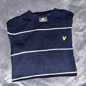 Randig marinblå tröja från Lyle & Scott - Snygg marinblå tröja från Lyle & Scott med tunna vita ränder och klassisk gul logga på bröstet. Tröjan har rund halsringning och långa ärmar, perfekt för en stilren och avslappnad look.