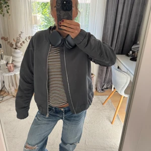Grå bomberjacka med dragkedja - Säljer en grå bomberjacka med klassisk passform och ribbade muddar. Jackan har en silkeslen yta, dragkedja framtill och lätt vaddering. Perfekt för dig som gillar en clean och stilren look. Snygg att bära öppen eller stängd.