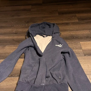 Mörkblå hoodie från Puma med dragkedja - Mörkblå hoodie från Puma med vit logga på bröstet. Tröjan har huva med dragsko, hel dragkedja framtill och två fickor. Insidan är mjuk och fleecefodrad för extra komfort. Perfekt för chill dagar eller när du vill ha en sportig look.