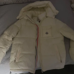 Vit Moncler pufferjacka med huva - Säljer en snygg vit pufferjacka från Moncler med avtagbar huva och klassisk logga på bröstet. Jackan har dragkedja med röd, vit och blå detalj, två fickor med dragkedja och en bröstficka. Perfekt för kalla dagar och riktigt stilren look.