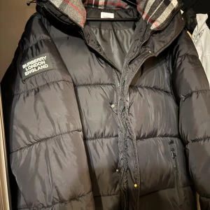 Svart dunväst från Burberry - Svart dunväst från Burberry med klassisk quiltad design och dragkedja framtill. Västen har en avtagbar huva med rutigt foder i grått, svart och rött. Märkets logga finns på bröstet. Perfekt för lager-på-lager och snygg till streetwear.