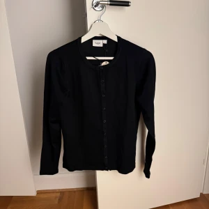 Svart kofta från Saint med knappar - En stilren svart kofta från Saint med rund halsringning och knäppning hela vägen framtill. Långärmad modell i mjukt material, perfekt att slänga på sig över en t-shirt eller linne. Enkel och clean look som funkar till det mesta.