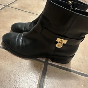 Svarta boots från Michael Kors - Snygga svarta boots i skinn från Michael Kors med guldfärgade detaljer och dragkedja baktill. Skorna har rund tå, platt sula och ett dekorativt MK-hänge på sidan. Perfekta för dig som vill ha en stilren och lyxig look till vardags.