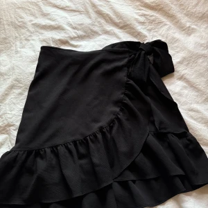 Svart omlottkjol med volang från Vero Moda - Svart kort omlottkjol från Vero Moda med volangkant och knyt i midjan. Kjolen har en snygg och enkel design som ger en trendig look. Perfekt för dig som gillar stilrena plagg med en twist.