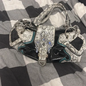 Svart och vit axelväska med ormmönster - Cool axelväska i svart och vitt med ormmönster, stora silvriga ringdetaljer och blingiga nitar och stenar framtill. Väskan har turkosa detaljer, flera fack och dragkedja baktill men även inuti. Materialet är glansigt syntet med skinnkänsla och väskan har en unik, edgy look.