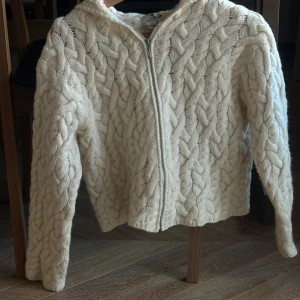 Vit stickad cardigan jacka med dragkedja - Stickad cardigan jacka med handgjord grovt flätmönster och dragkedja framtill. Jackan har lång ärm och huva. Passar S. 