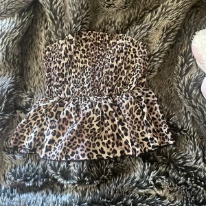 Leopardmönstrad tubtopp från Romwe - Säljer en tubtopp från Romwe i storlek S med leopardmönster. Toppen har smockad överdel som sitter tight och en volang nedtill som ger en snygg siluett. Färgerna är beige, brun och svart. Perfekt för dig som vill sticka ut med ett djuriskt print.