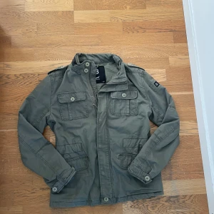 Olivgrön jacka från Brandit, storlek M - Snygg olivgrön jacka från Brandit i storlek M. Jackan har fyra stora fickor framtill med lock och knappar, axelklaffar och hög krage. Tillverkad i bomull och stängs med dragkedja och knappar. Perfekt för dig som gillar en stilren och funktionell look.