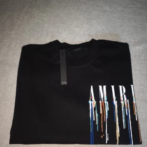 Svart Amiri T-shirt - Svart t-shirt från AMIRI med stort vitt logotryck och färgstänk.                         Tillverkad i 100% bommul i storlek S, oberzised. Äkta ny med lapp, passar s/m
