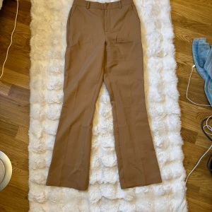 Beiga/bruna kostymbyxor från Bik Bok - Snygga beiga/bruna kostymbyxor från Bik Bok i klassisk modell med raka ben och hällor för bälte. Byxorna har diskreta fickor fram och bak, och är tillverkade i ett mjukt material som känns skönt mot huden. Perfekta för en clean och stilren look.