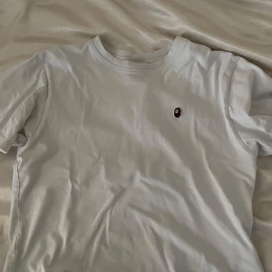 Vit BAPE t-shirt med ape-logga - Vit t-shirt från BAPE i bomull med klassisk ape-logga broderad på bröstet. Enkel och clean design med rund hals och korta ärmar. Perfekt för dig som gillar streetwear och vill ha något stilrent men ändå ikoniskt. Tröjan är äkta ✅