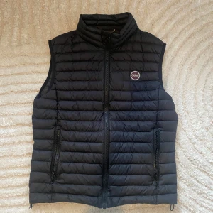 Svart dunväst från Colmar, strl 50 - Svart dunväst från Colmar i bra skick med horisontella quiltade paneler och hög krage. Västen har två sidofickor med dragkedja och en innerficka, samt Colmar-logga på bröstet. Perfekt för lager-på-lager och har en stilren, sportig look.