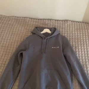 Grå hoodie från McKenzie med tryck - Säljer en grå hoodie från McKenzie med vit logga framtill och stort tryck på ryggen. Tröjan har huva med snörning och en klassisk känguruficka. Perfekt för chill dagar eller när du vill ha en enkel streetstyle-look.