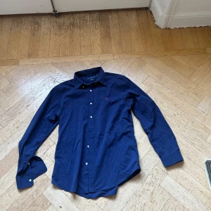 Mörkblå skjorta från Ralph Lauren - Snygg mörkblå skjorta från Ralph Lauren i slim fit med klassisk krage och knappar framtill. Skjortan har det ikoniska röda Polo-logot broderat på bröstet och är tillverkad i mjuk bomull. Perfekt för dig som gillar stilrena och tidlösa plagg.
