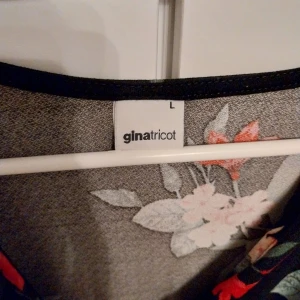 Blommig omlottopp från Gina Tricot - Snygg svart omlottopp från Gina Tricot med blommigt mönster i rött, beige och grönt. Toppen har v-ringning, långa ärmar och knyt i midjan. Perfekt för dig som vill ha en feminin och trendig look med lite extra detaljer. Storlek:L (Large)
