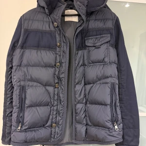 Mörkblå Moncler pufferjacka med huva - Snygg mörkblå pufferjacka från Moncler med huva, dragkedjor på fickorna och knappar framtill. Jackan har quiltad design, Moncler-logga på ärmen och kombinerar glansigt och matt material för en modern look.