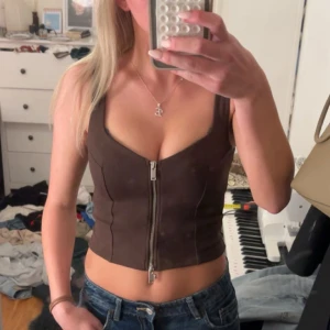 Brun Zara -topp med dragkedja - Snygg brun Zara -topp med breda axelband och en rejäl silverfärgad dragkedja framtill. Toppen är croppad och har en figurnära passform som framhäver midjan. Materialet känns mjukt och stretchigt, troligen bomull med lite syntet. Perfekt för dig som gillar trendiga och edgy plagg.