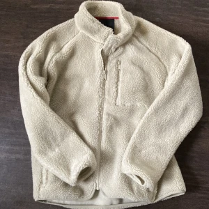 Beige teddyjacka från Tretorn - Mysig beige teddyjacka från Tretorn, modell Sarek Camp Jacket W's. Jackan har hel dragkedja framtill, hög krage och en praktisk bröstficka med dragkedja. Tillverkad i mjuk fleece som håller dig varm under kyliga dagar. Perfekt för dig som gillar chill och outdoor.