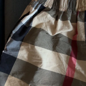 Rutig shorts med resårmidja - Snygga shorts med klassiskt rutigt mönster i beige, svart, grått och en röd detalj. De har en bred resår i midjan för skön passform och är tillverkade i ett lätt bomullstyg. Perfekta för varma dagar när du vill ha en chill och stilren look.