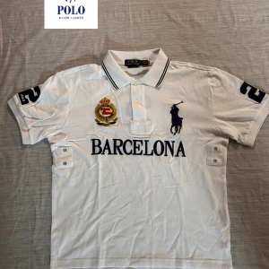 Vit pikétröja Polo Ralph Lauren L - Vit pikétröja från Polo Ralph Lauren i slim fit med broderad logga, emblem och texten BARCELONA på bröstet. Tröjan har klassisk krage med knappar, svarta detaljer på kragen och ärmarna samt stora siffror på ärmarna. Perfekt för dig som gillar sportig och stilren look.