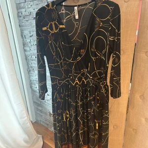 Svart omlottklänning Chiara Forni - Svart långklänning från Chiara Forni Milano med guldigt kedjemönster, omlottmodell och trekvartsärm. Klänningen har knyt i midjan och dekorativa ränder i rött och grönt längst ner. Snygg och elegant look med v-ringad hals.