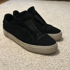 Svarta sneakers från Axel Arigato - Snygga svarta sneakers från Axel Arigato med minimalistisk design och vit platt sula. Skorna är i mocka med elastisk detalj över vristen och har diskret guldfärgad logga på sidan. Perfekta för dig som gillar stilrena och trendiga skor.