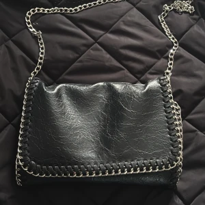 Svart axelväska med kedja och skinn - Snygg svart axelväska i skinnimitation med silverfärgad kedja som axelrem. Väskan har dekorativa kedjedetaljer längs kanterna och ett innerfack med dragkedja.
